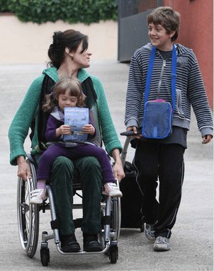 Mujer en silla con su hija pequeña en las piernas y charlando con el hijo mayor a la salida del colegio.